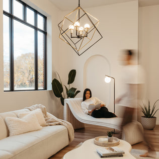 A modern living room with large windows features the Artika Peyton LED modern mid-century pendant light, neutral furnishings, and cozy decor. A woman reads on a chaise lounge as a blurred figure walks by, with plants adding warmth to the space.