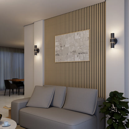 A modern living room features a gray sofa, wooden slat accent wall, abstract artwork, potted plant, and two Artika Dani modern LED indoor-outdoor wall lights (5 CCT), with a dining area visible in the background.