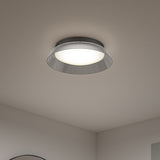 A modern ceiling light fixture with a wide, metallic, slightly flared shade and a central glowing bulb, mounted on a white ceiling in a minimal, neutral-colored room.