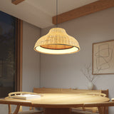 A woven rattan pendant light hangs above a round wooden dining table with matching chairs in a minimalist room. A simple artwork and a vase with branches decorate the light-colored wall.