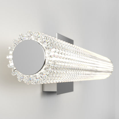 The Artika Exalto LED bathroom vanity light features a modern cylindrical design with multiple rows of clear crystal accents on a sleek rectangular metal base, casting bright white light against a plain, light-colored wall.