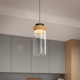 A modern pendant light with a cylindrical glass shade and a wooden accent hangs from the ceiling in a contemporary kitchen with gray cabinets and wood details.