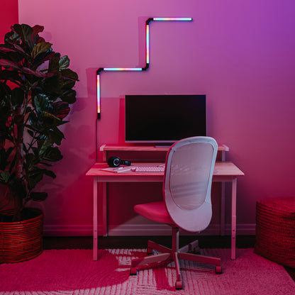 A modern workspace features a computer on a wooden desk, a white mesh office chair, headphones, a potted plant, woven basket, and the Artika Fitch smart foldable RGB LED gaming floor lamp illuminating the room with pink and purple lighting.