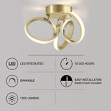 Modern gold ceiling light with three LED rings, shown mounted on a grey background. Feature icons below list: LED integrated, 50,000 hours, dimmable, 1300 lumens, and easy installation with safety hook included.