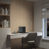 A modern home office features a gray chair at a white desk with a closed laptop, notebook, and coffee mug. Wooden slats decorate the wall, and shelves with books are on the left. A window with blinds is on the right.