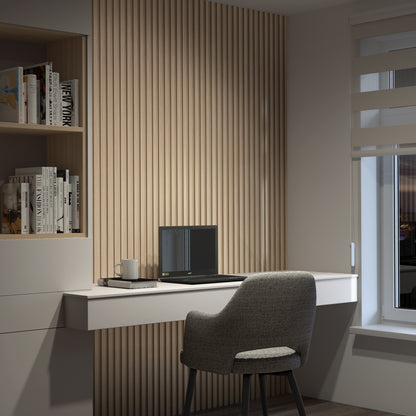 A modern home office with a gray chair, white floating desk, closed laptop, mug, and notepad. The wall features Artika's Panolok 30.67 ft wall panel kit (pack of 8), shelves with books, and a window with beige blinds showing evening light.