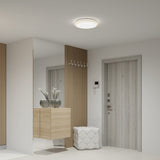 A modern hallway with light wood and white decor, featuring a light wood door, a floating cabinet with two glass vases of white flowers, a quilted white ottoman, and a wall mirror.
