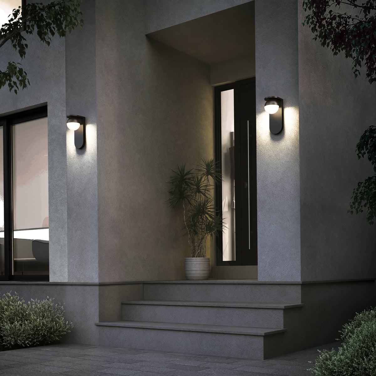 Modern house entrance at dusk with two wall-mounted lights, a black door, potted plant, gray steps, and surrounding greenery. The exterior walls are gray and the area is softly illuminated.