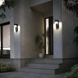 Modern house entrance at dusk with two wall-mounted lights, a black door, potted plant, gray steps, and surrounding greenery. The exterior walls are gray and the area is softly illuminated.