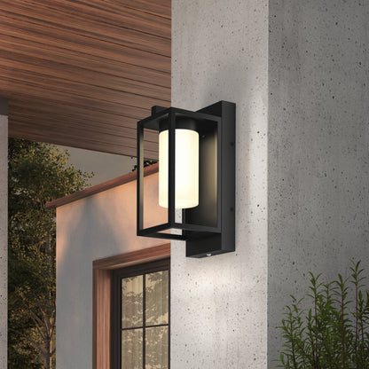 The Artika Aurel Outdoor LED Light, 3CCT is a modern black metal wall sconce with a cylindrical white fixture, mounted on a concrete wall next to a house with plants and trees in the background.