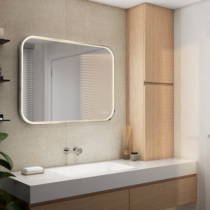 A modern bathroom with a large, illuminated rectangular mirror above a white sink, light wood cabinets, a wall-mounted faucet, and a small plant on the left side of the countertop.