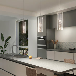 Modern kitchen with gray cabinets, a long island with sink, and three Artika Bubble Flow 1-light pendant 2 lights above. A window reveals greenery outside; a single apple, notepad on the counter, and decor by the backsplash complete the scene.
