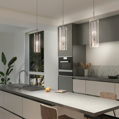 Modern kitchen with gray cabinets, a long island with sink, and three Artika Bubble Flow 1-light pendant 2 lights above. A window reveals greenery outside; a single apple, notepad on the counter, and decor by the backsplash complete the scene.
