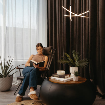In a cozy, well-lit room featuring the Artika Orly modern LED pendant light 5 CCT, a woman in jeans, a sleeveless top, and slippers reads by the window near potted plants, books, a candle, and a round coffee table.