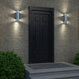 A black paneled door with a handle is set in a brick wall, flanked by two modern wall lights. There are green bushes on either side of the door, and three steps lead up to the entrance.