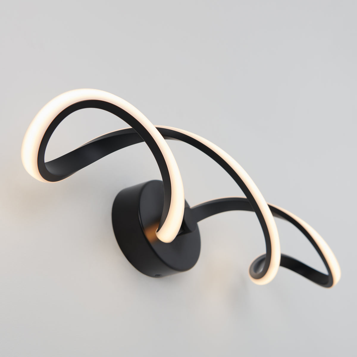 A modern wall light with two curved, looping LED strips attached to a black circular base, mounted on a white wall. The design is minimalist and artistic, emitting a soft, warm glow.