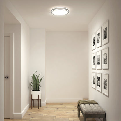 A minimalist hallway with light wood floors, white walls, the Artika Altitude 15 in. LED modern flush mount light, a potted plant on a stand, a cushioned bench with a blanket, and a grid of framed photos on the wall.