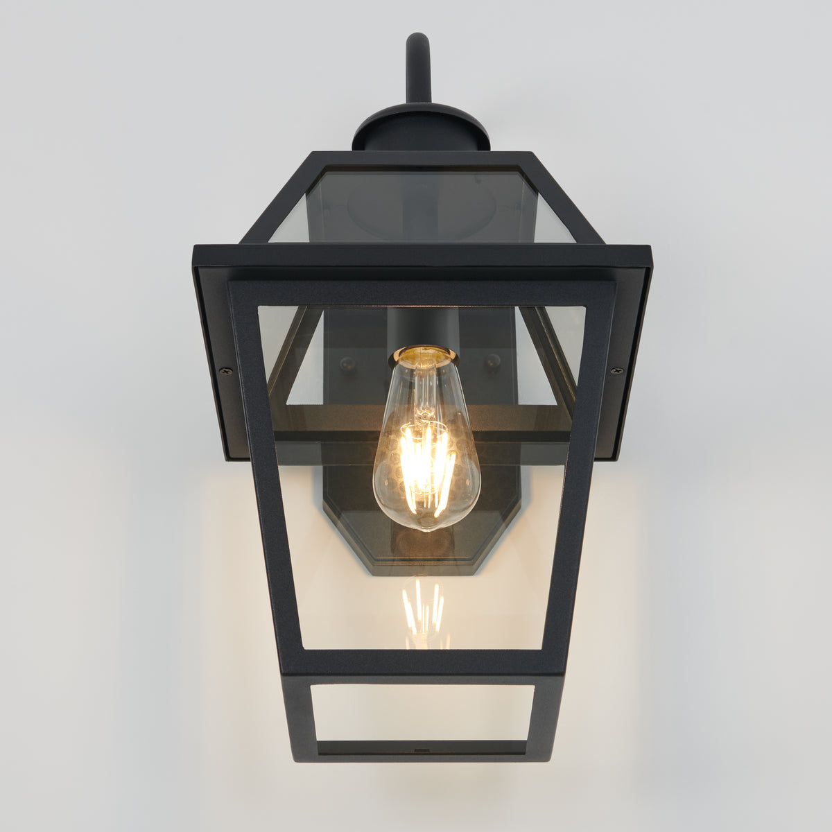 A black metal and glass wall lantern with a vintage-style exposed filament bulb, mounted on a white wall. The lantern has a rectangular, tapered design with clear glass panels.