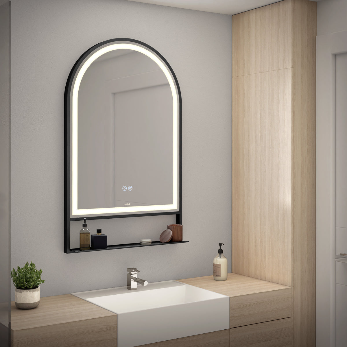 A modern bathroom with a large, arched, backlit mirror above a white sink. There is a wooden cabinet, a soap dispenser, potted plant, and small items on a glass shelf below the mirror.