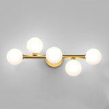 A modern wall sconce with five round white glass globes set on a horizontal gold metal bar, mounted on a gold circular base against a light gray wall.