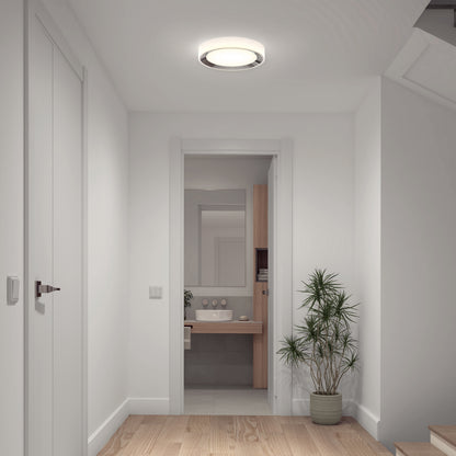 A bright, modern hallway with white walls, wooden floor, a potted plant, and a view into a bathroom with large mirror and sink is illuminated by the Artika Cooper LED flush mount light with remote.