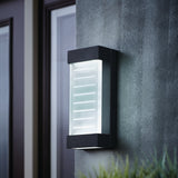 A modern rectangular outdoor wall light fixture with frosted glass and horizontal lines is mounted on a textured gray wall next to a door and window. The light is turned on, emitting a soft glow.