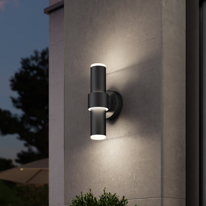 The Artika Dani modern LED indoor-outdoor wall light 5 CCT, a sleek black cylinder fixture, casts up-and-down lighting on a gray exterior wall at dusk, with blurred trees and a patio umbrella in the background.