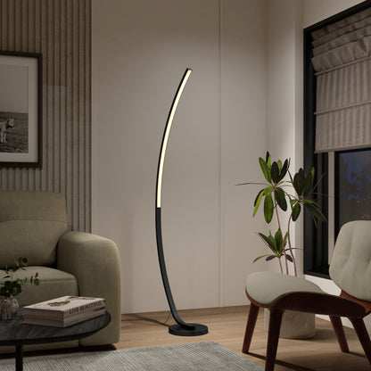 A modern living room includes Artika’s Arc LED Floor Lamp in black, casting soft light beside a green sofa, a plant, and a wooden chair, with a large window and framed wall art visible in the background.