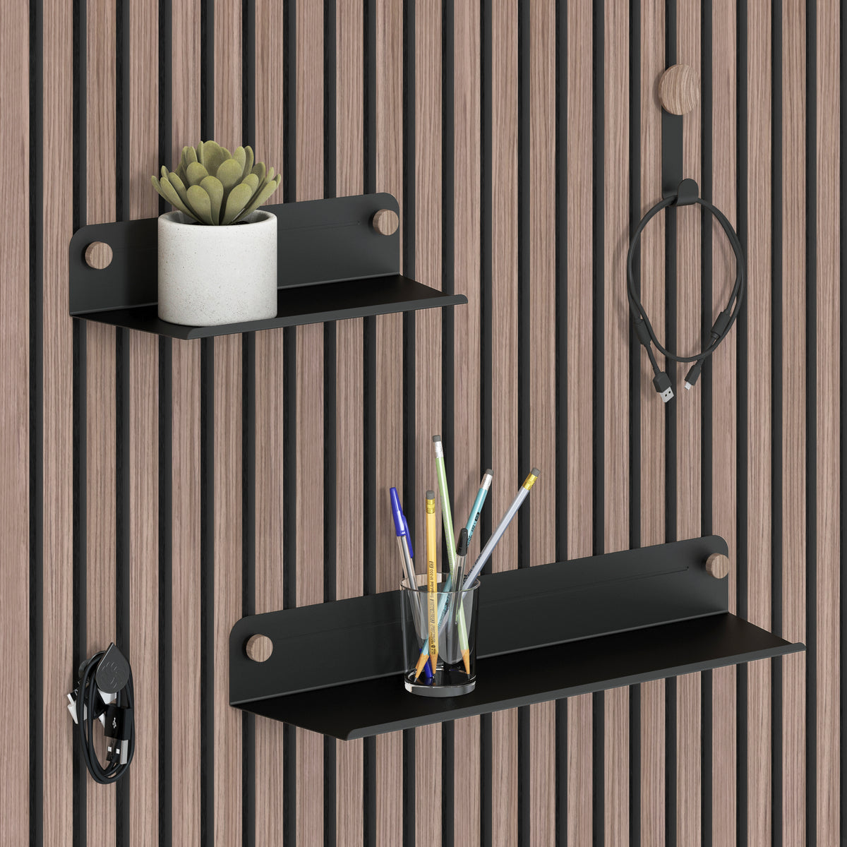 Two black metal shelves are mounted on a wooden slat wall. The top shelf holds a small potted succulent, and the lower shelf holds a clear cup with pens. Nearby hooks hold a cable and a pair of headphones.