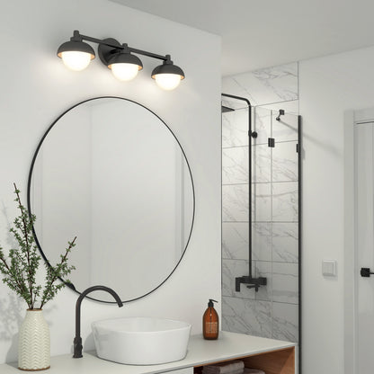 A modern bathroom featuring Artika’s Boden LED mid-century vanity light, a round mirror, black faucet and sink, white vase with green branches, soap dispenser, and a glass-enclosed marble shower with black fixtures.