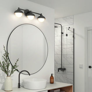 Modern bathroom with a round mirror, white vessel sink, black faucet, and a vase with greenery. Above the mirror is the Artika Boden 27-in Integrated LED 5-CCT Vanity Light Bar. In the background, a shower features white marble tiles and black fixtures.