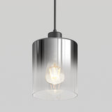 A modern pendant light with a clear cylindrical glass shade and a visible glowing bulb, suspended from a black cord against a plain light gray background.