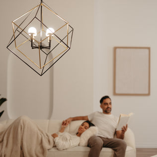 An Artika Peyton LED modern mid-century pendant light hangs in the foreground, while a couple relaxes on a cream-colored sofa in the background; one reads a book, the other lounges under a blanket.