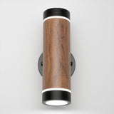 A modern cylindrical wall sconce with a wood grain finish, black metal end caps, and circular white LED lights on both ends, mounted on a round black wall plate against a plain white background.