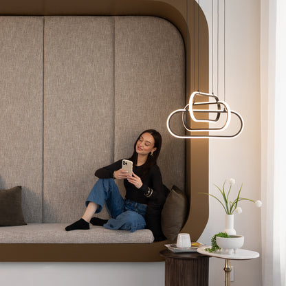 A woman sits comfortably on a modern bench with gray upholstery, smiling at her smartphone. Above, the Artika Lewis modern LED pendant light 3 CCT hangs like decorative clouds. Nearby is a small table with flowers and a cup.