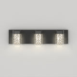 A modern wall sconce with three lights, each featuring a cylindrical glass shade and a black rectangular backplate, mounted on a light gray wall. The lights are lit, emitting a soft glow.
