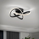 A modern ceiling light fixture with intertwined black bands and integrated LED lights, mounted on a white ceiling in a minimalist room. A plant and curtains are partially visible in the lower corners.
