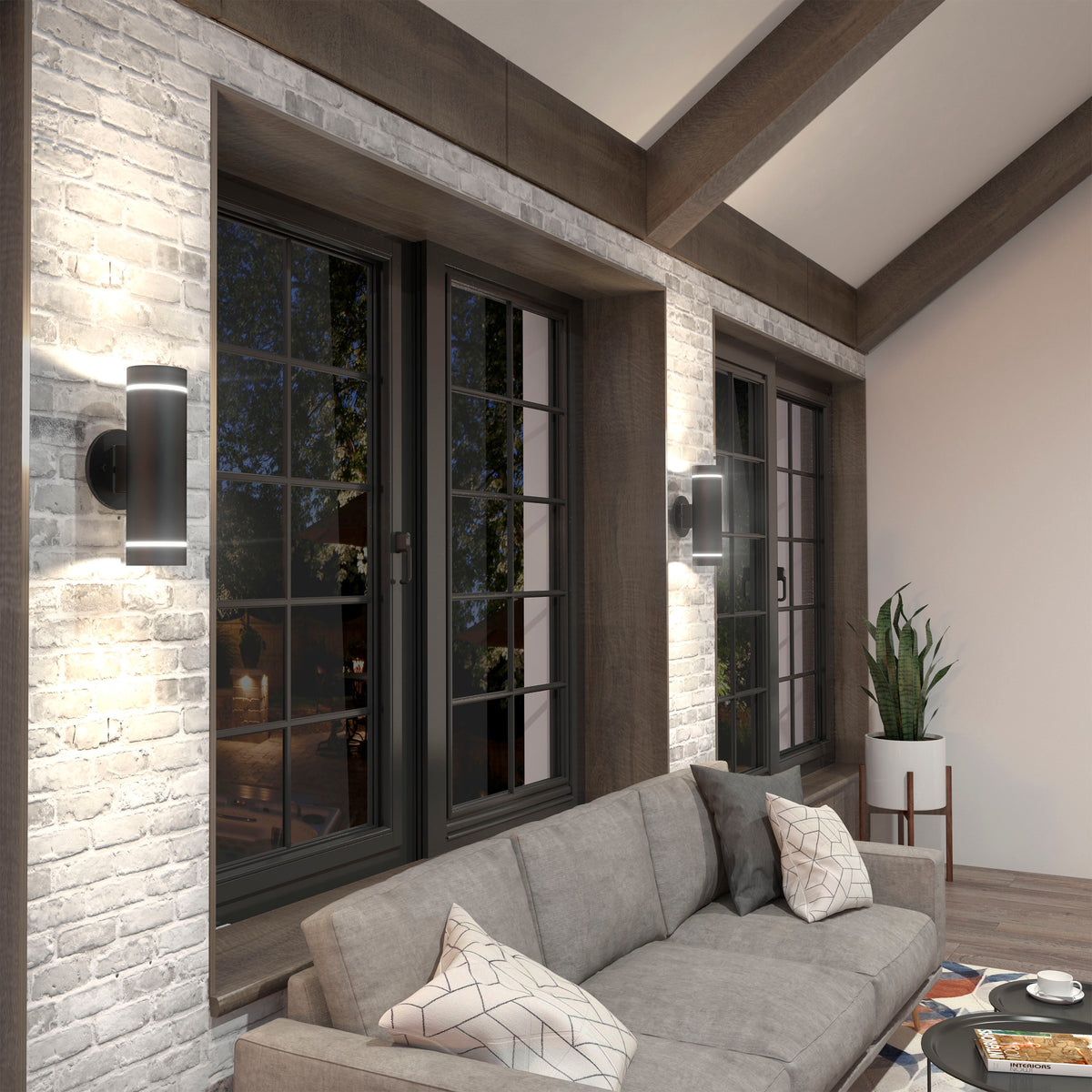 A modern living room with a gray sectional sofa, patterned pillows, large black-framed windows, wall sconces on white brick, a potted plant in the corner, and wooden ceiling beams.