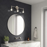 A modern bathroom with a round mirror, marble sink, wall-mounted faucet, three-bulb vanity light, potted plant, soap dispenser, towel ring with a gray towel, and a window with white blinds.