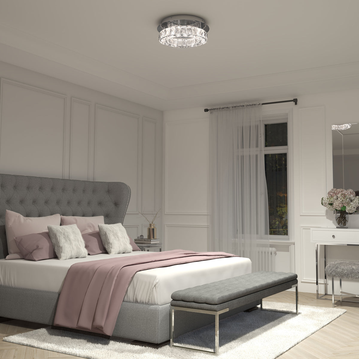 A modern bedroom with a gray upholstered bed, white and pink bedding, a gray bench at the foot, a vanity with flowers, white paneled walls, sheer curtains, and a crystal ceiling light.