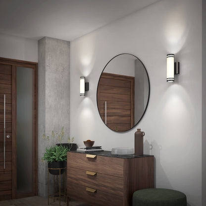 A modern entryway with a round mirror above a wooden dresser, potted plant, decorative items, and two wall sconces casting soft light on white walls near a wooden door.