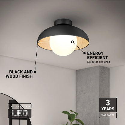 The Artika Boden LED flush mount light 5 CCT boasts a sleek black and wood finish, integrated LED for energy efficiency, and a 3-year warranty. Shown in a minimalist room with a gray wall and green plant.