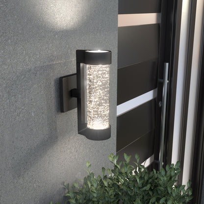 The Artika Bubble Element LED indoor-outdoor light features a modern cylindrical design with a black frame and textured glass, shown illuminated on a gray wall by a metal gate above green leafy plants.