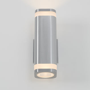 The Artika Mettle LED indoor-outdoor wall light is a modern, cylindrical stainless steel sconce with a brushed finish that shines soft white light from top and bottom, stylishly mounted on a plain light gray wall for energy-efficient illumination.