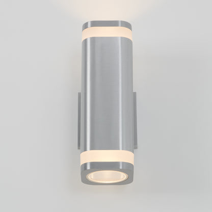 The Artika Mettle LED indoor-outdoor wall light is a modern, cylindrical stainless steel sconce with a brushed finish that shines soft white light from top and bottom, stylishly mounted on a plain light gray wall for energy-efficient illumination.