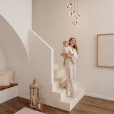 A woman holding a baby stands on modern white stairs in a stylish, minimalistic room with soft lighting, wooden floor, and neutral decor, including a bench with cushions and large lanterns.
