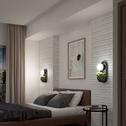 A modern bedroom features a brown bed, gray and white bedding, white brick accent wall, abstract art, plants, beige curtains, a side table, and Artikas Bloomfield LED modern mid-century wall sconce (5 CCT).