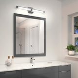 A modern bathroom features a large square mirror with a grey frame, a sleek vanity with drawers, a faucet, toiletries, and a potted plant on the windowsill. A wall light is mounted above the mirror.