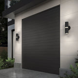 A modern house exterior with a large, dark gray garage door and two cylindrical wall lights on either side, mounted on light-colored walls. Green plants are visible near the entrance.
