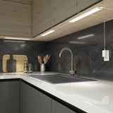 A modern kitchen countertop with a sleek sink, spring faucet, cutting boards, glass cups, and utensils holder. The backsplash features dark, chevron-patterned tiles and under-cabinet lighting.